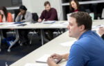A group of adults sit at tables in a classroom, taking notes and listening during a discussion.