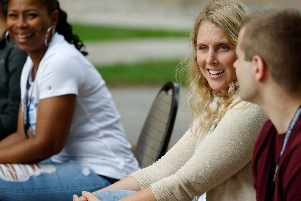 Three people sit outdoors, talking and smiling. The woman in the center faces a man, engaging in conversation, while another woman sits nearby, smiling.