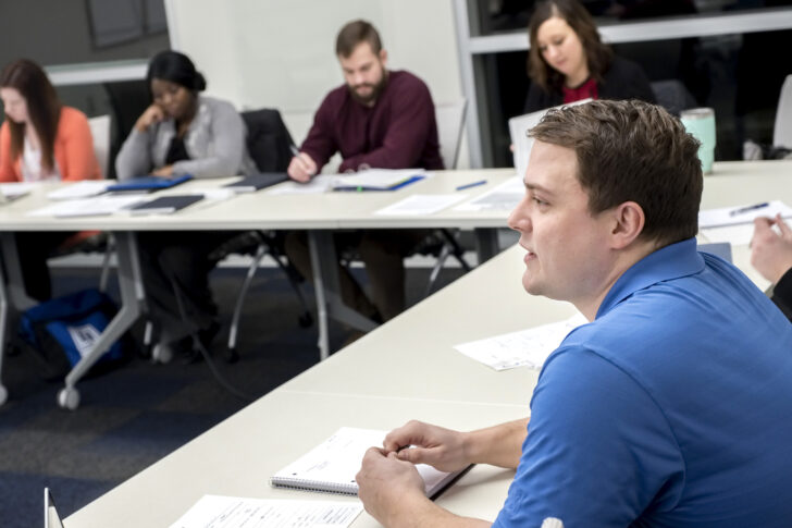A group of adults sit at tables in a classroom, taking notes and listening during a discussion.