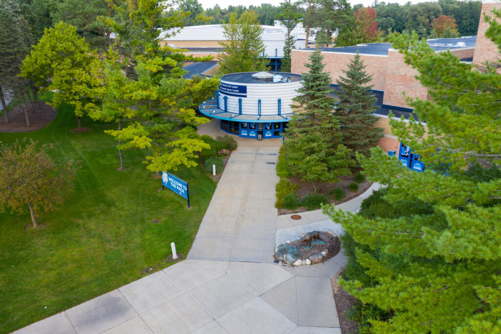 Aerial view of a building entrance surrounded by trees, with blue doors and a sign that reads “Fieldhouse” on a grassy campus walkway.
