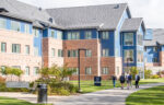 Four people walk on a pathway outside a modern brick and blue building, surrounded by grass and shrubs on a sunny day.