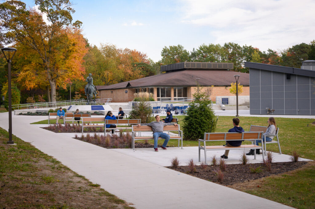People sit on benches and walk along a concrete path in a landscaped outdoor campus area with trees, a statue, and modern buildings in the background.