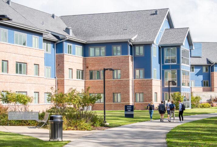 Four people walk on a pathway outside a modern brick and blue building, surrounded by grass and shrubs on a sunny day.