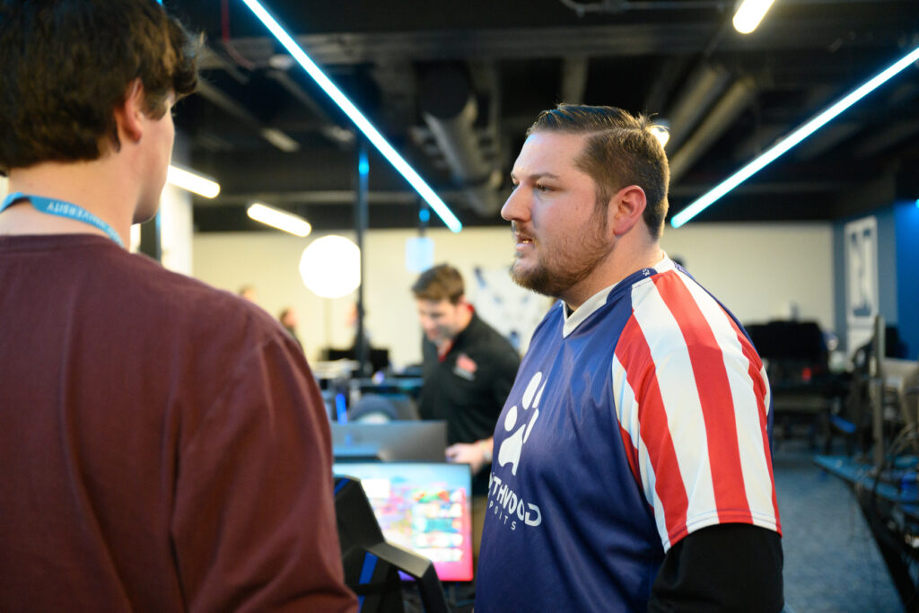Two people are talking in a room with computers; one person is wearing a striped sports jersey. Other individuals are working in the background.