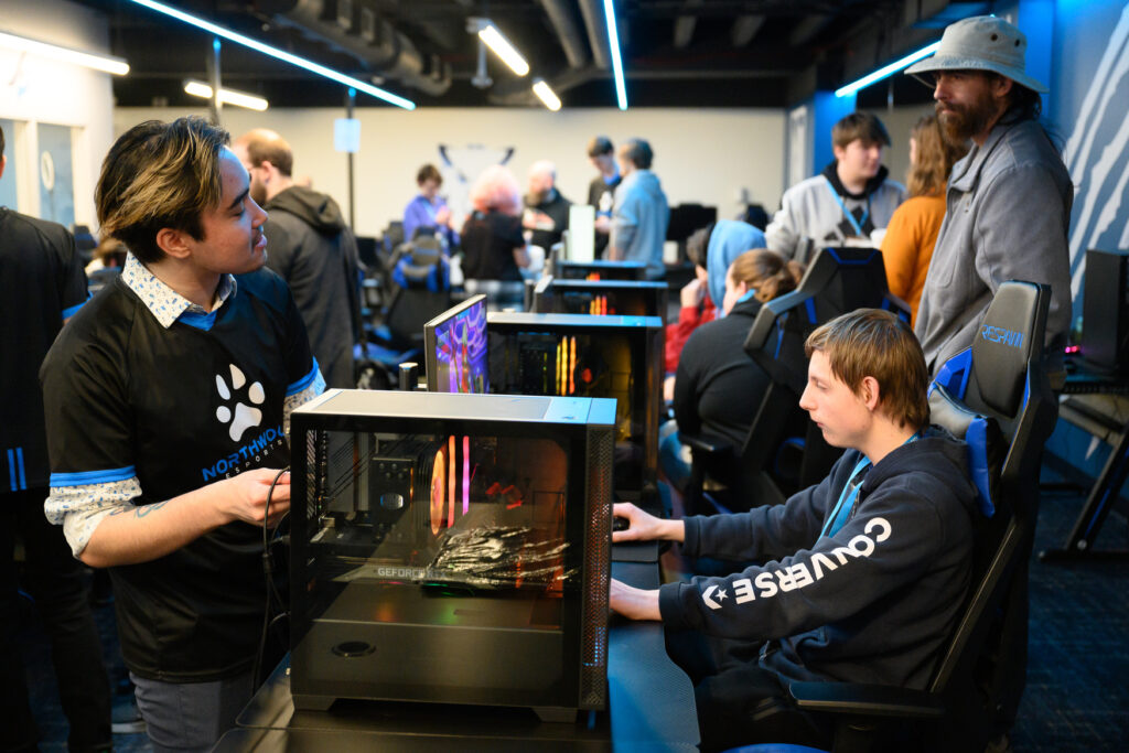 Several people are gathered in a gaming room; one person sits at a computer playing a game while others stand and observe or interact in the background.