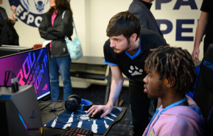 Two men focus on a computer screen in a gaming setup, one sitting and using the keyboard while the other stands and points at the monitor; other people are in the background.