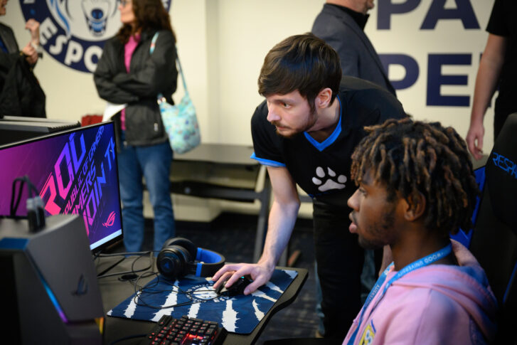 Two men focus on a computer screen in a gaming setup, one sitting and using the keyboard while the other stands and points at the monitor; other people are in the background.