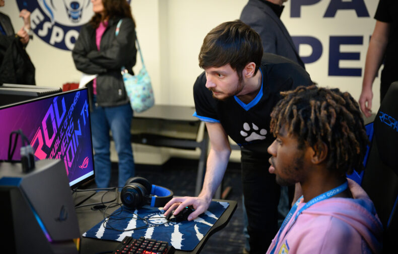 Two men focus on a computer screen in a gaming setup, one sitting and using the keyboard while the other stands and points at the monitor; other people are in the background.