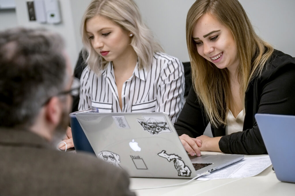 Two women work at a table with laptops and papers, while a man sits across from them. The laptops have visible stickers on their covers, suggesting they could be collaborating on projects related to online business degree programs and exploring various business career options.