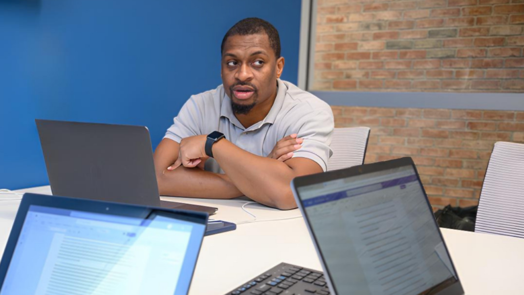 A man sits at a conference table with a laptop, speaking, while two open laptops with documents are visible in the foreground.