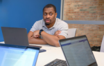 A man sits at a conference table with a laptop, speaking, while two open laptops with documents are visible in the foreground.