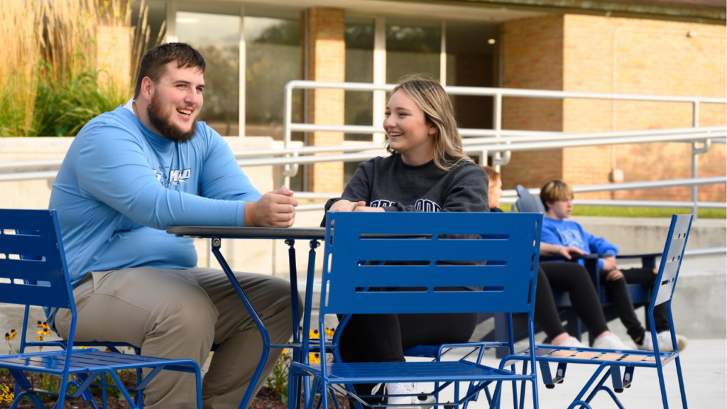 Two people sit at a blue metal table outdoors, talking and smiling—perhaps sharing college visit tips as they enjoy the sunny afternoon. Two others sit on benches in the background near a building with large windows, reflecting the welcoming atmosphere of a successful campus visit.